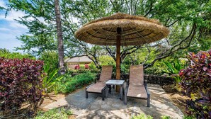 An outdoor seating area with a thatched umbrella, stone flooring, and lush greenery. - Beautiful Townhouse - Great Amenities and Spacious Lanai! Comes With Beach Pass! - Vrkcv (Kamuela)