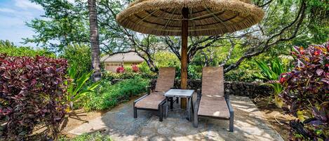 An outdoor seating area with a thatched umbrella, stone flooring, and lush greenery.