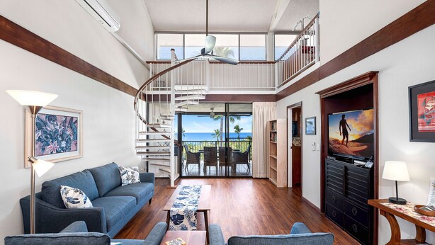 A spacious living room with a blue sofa, a wooden coffee table, and a TV stand. A spiral staircase leads to a balcony with a view of the ocean. - Ocean & Golf Course Views - 2 Bdrm With Loft - AC - Private Lanai -vrkcv (Kailua-Kona)