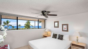 A bedroom with a large bed, two bedside tables with lamps, a ceiling fan, a window with a view of the ocean, and two framed pictures on the wall. - Ocean & Golf Course Views - 2 Bdrm With Loft - AC - Private Lanai -vrkcv (Kailua-Kona)