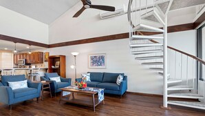 A spacious living room with a blue sofa set, a coffee table with a floral centerpiece, and a white spiral staircase. - Ocean & Golf Course Views - 2 Bdrm With Loft - AC - Private Lanai -vrkcv (Kailua-Kona)