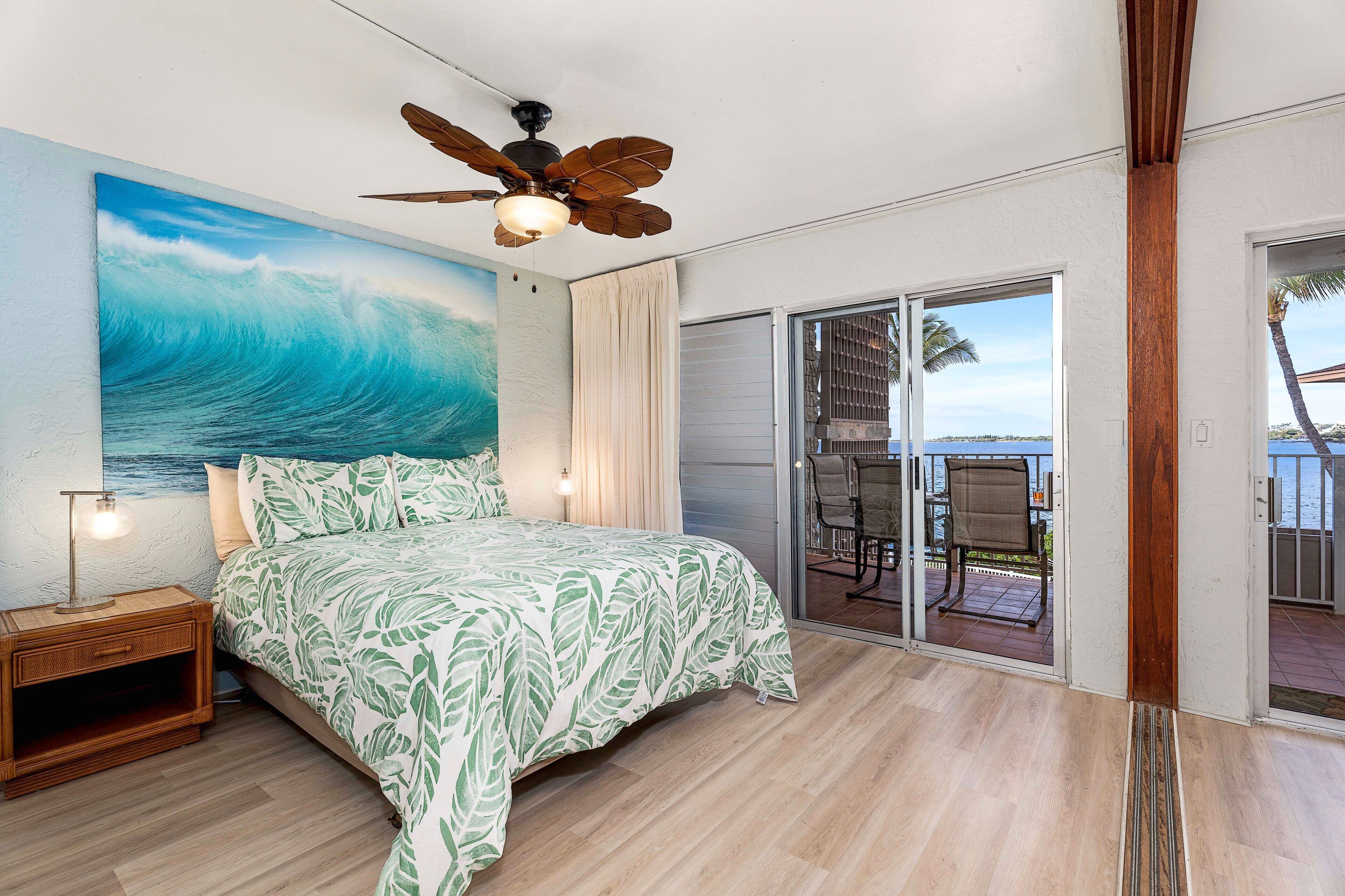 A bedroom with a large ocean wave painting, a ceiling fan, a bed with a leaf-patterned bedspread, a nightstand, and a view of a balcony with a chair.