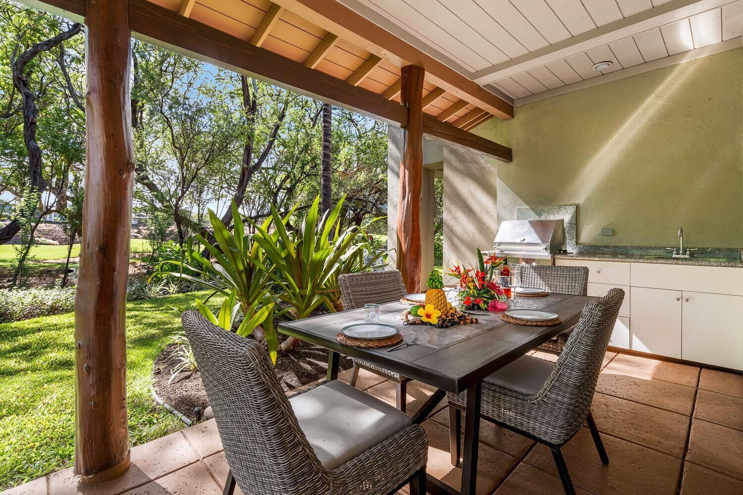 An outdoor patio with a table set for dining, surrounded by wicker chairs, and a kitchen area with a grill.
