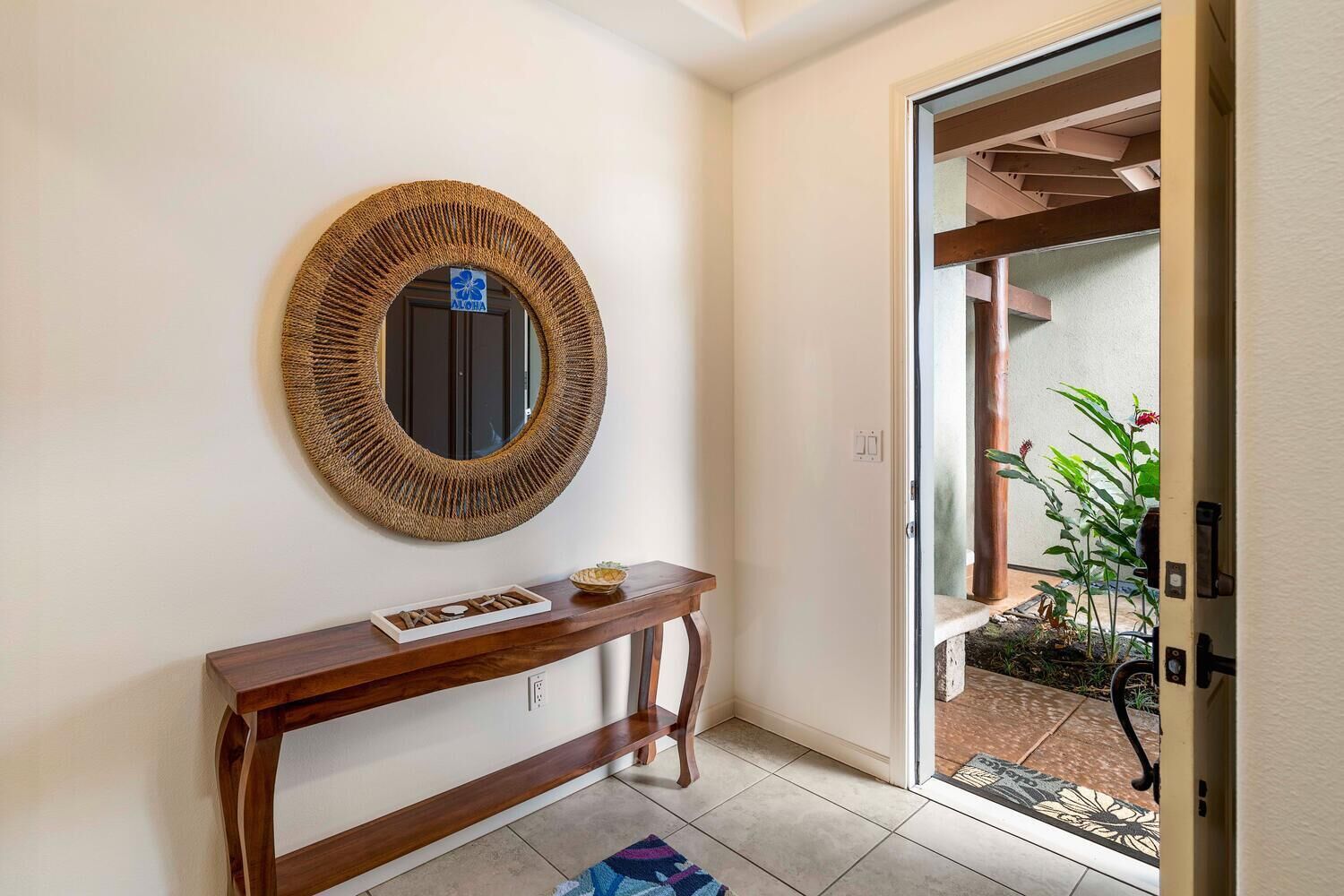 Un pasillo con un espejo tejido, una consola de madera y una puerta que da a un balcón con plantas.