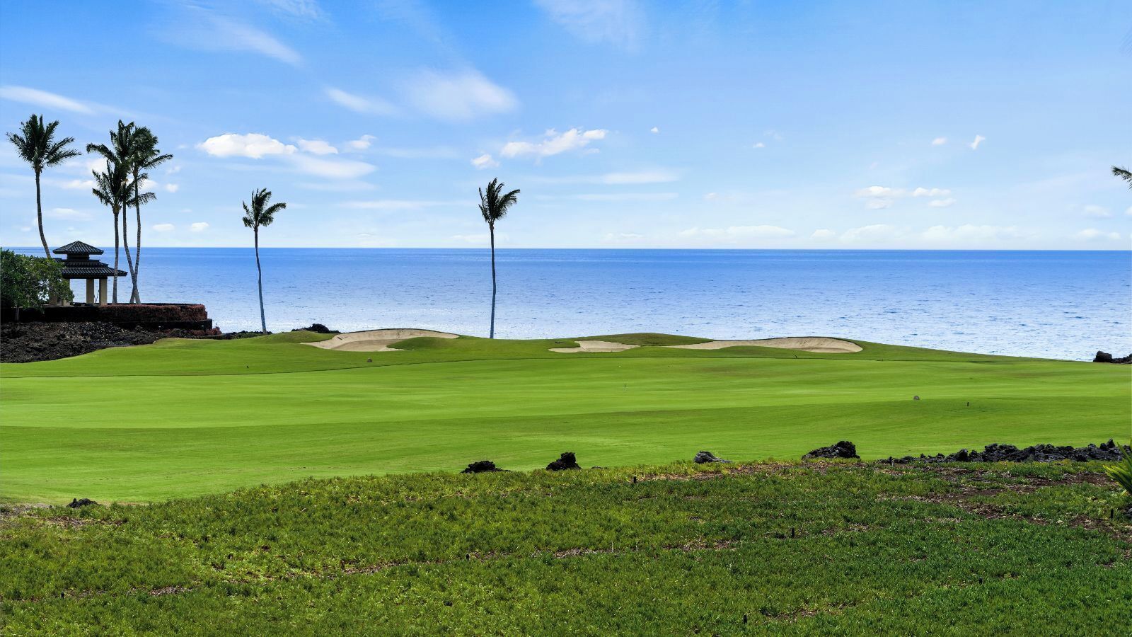 A golf course with a view of the ocean, palm trees, and a small pavilion.