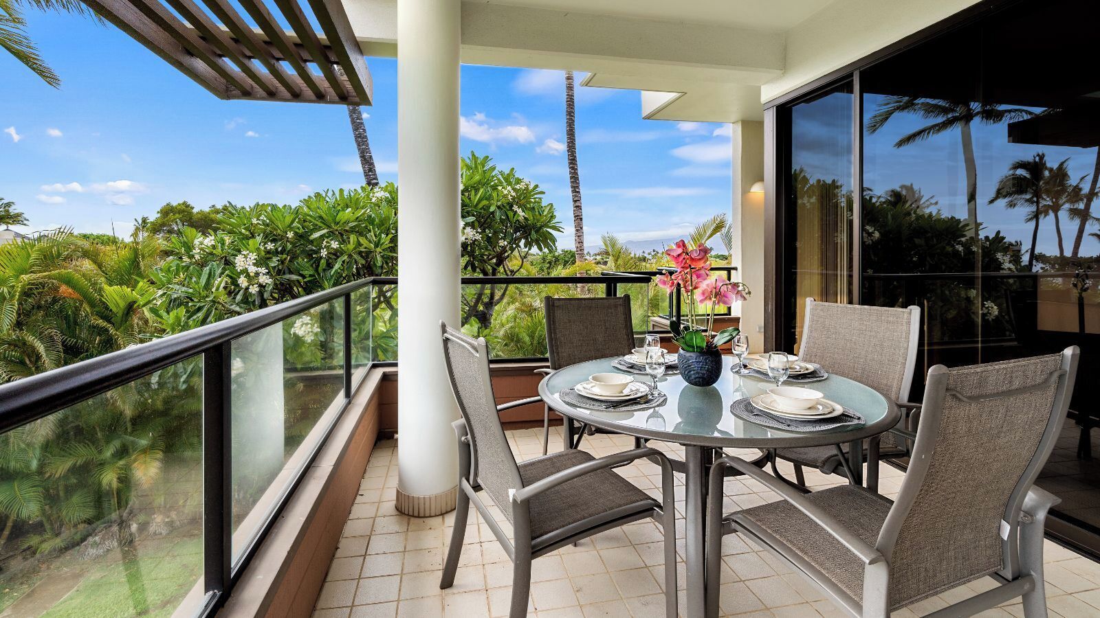 A balcony with a glass railing, a round table set for dining, and chairs.