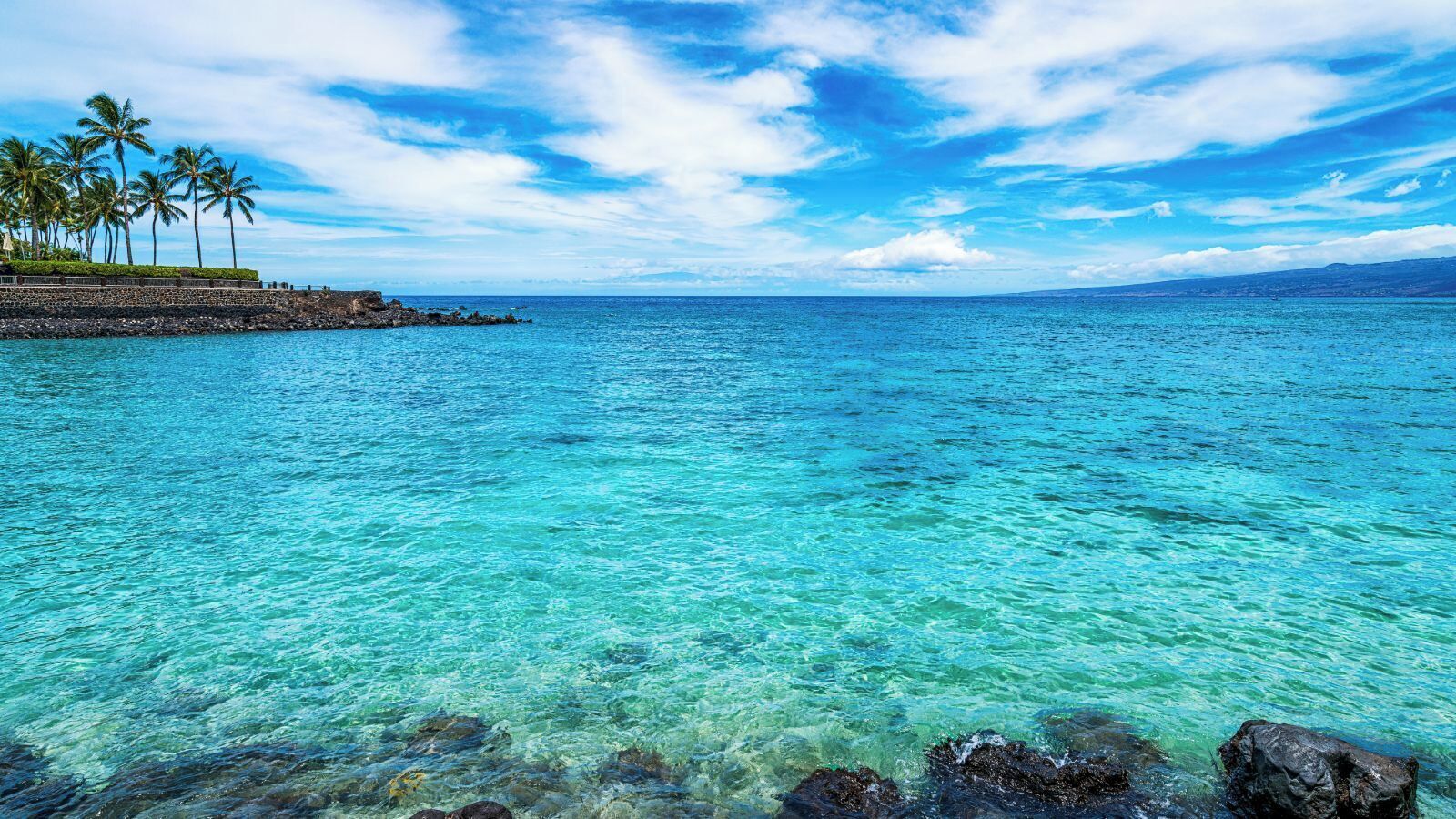 Mar azul transparente con rocas visibles en la orilla y una costa rocosa distante.