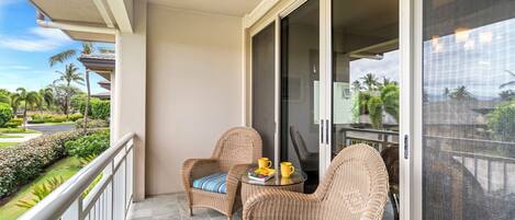 A balcony with wicker chairs and a small table, overlooking a landscaped garden and palm trees.