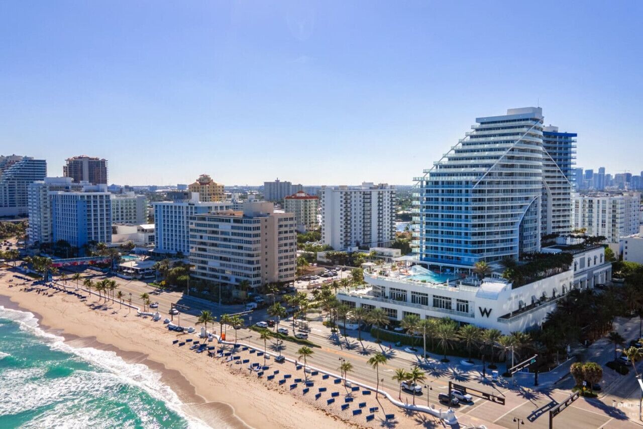 Un paysage urbain côtier avec un hôtel imposant à la forme incurvée, doté de balcons, une plage avec des transats et une vue dégagée sur l’océan.