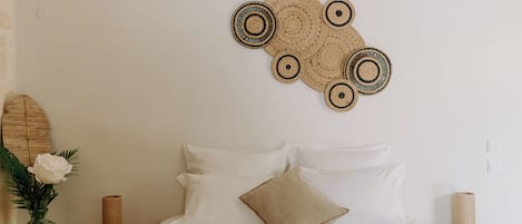 A bedroom with a bed, white pillows, a beige throw, a wall hanging, a vase with flowers, and a wooden nightstand with a candle.