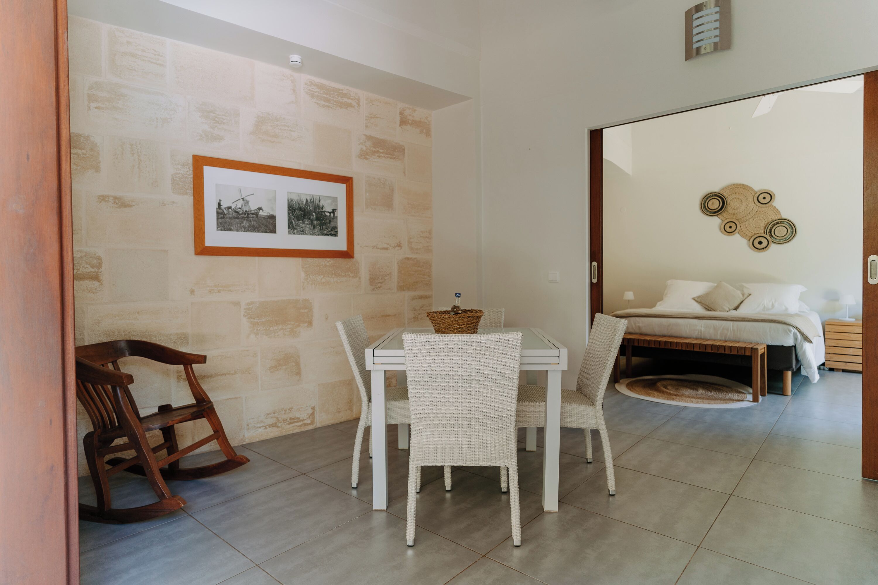 A dining area with a table and chairs, a rocking chair, a bed with a headboard, and a wall-mounted picture frame.