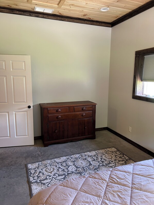 Un dormitorio con techo de madera, una puerta blanca, un aparador de madera oscura, una alfombra estampada y una cama con colcha blanca.