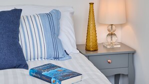 Una cama bien hecha con un libro titulado "Odyssey" de Stephen Fry, una lámpara y un jarrón decorativo sobre una mesita de noche.