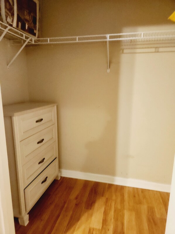 Una habitación pequeña y vacía con piso de madera, un aparador blanco con cuatro cajones y un armario blanco con barra para colgar.