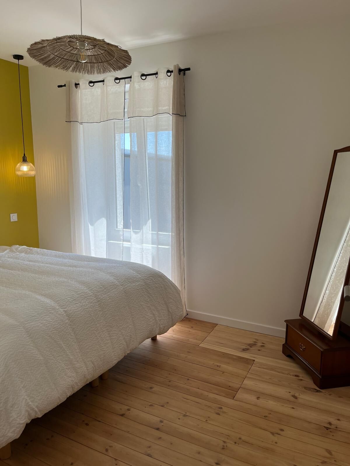 Une chambre à coucher avec un lit, un plancher en bois, un luminaire suspendu, un miroir et une fenêtre avec des rideaux légers.