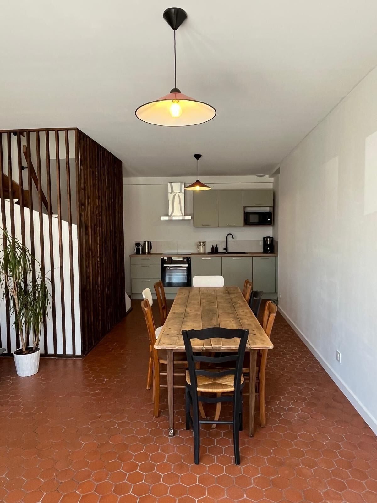 A modern kitchen with a dining area, featuring a wooden table and chairs, a hanging light fixture, and a potted plant.
