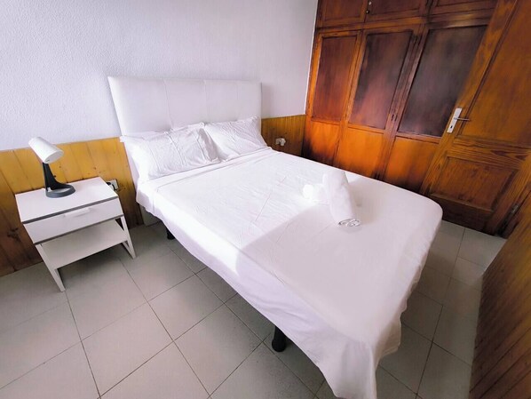 Habitación individual con una cómoda de madera, una mesita de noche con lámpara y una colcha blanca.