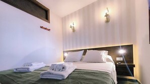 A neatly made bed with white linens and a green throw blanket, a bedside table with a lamp, and a wall-mounted light fixture. - Atlantic Dream Sunset (Mesa del Mar)