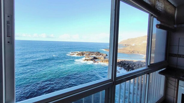A room with a large window overlooking a rocky coastline and the sea. - Atlantic Dream Sunset (Mesa del Mar)
