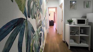 Un pasillo con un gran mural de hojas tropicales, un estante blanco con microondas, y una puerta que da a otra habitación.