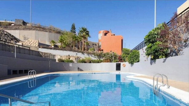 A clear blue swimming pool with steps leading into the water, surrounded by a low wall and a hillside with vegetation. - Argos Sunset Apartment (Candelaria)