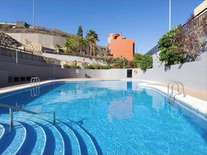 A clear blue swimming pool with steps leading into the water, surrounded by a low wall and a hillside with vegetation. - Argos Sunset Apartment (Candelaria)