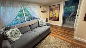 A cozy living room with a grey sofa, patterned cushions, a large window with sheer curtains, a wooden floor, and a poster on the wall.