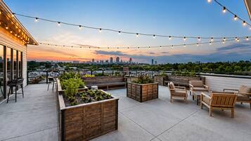 A rooftop patio with wooden planters, outdoor furniture, and string lights against a city skyline.