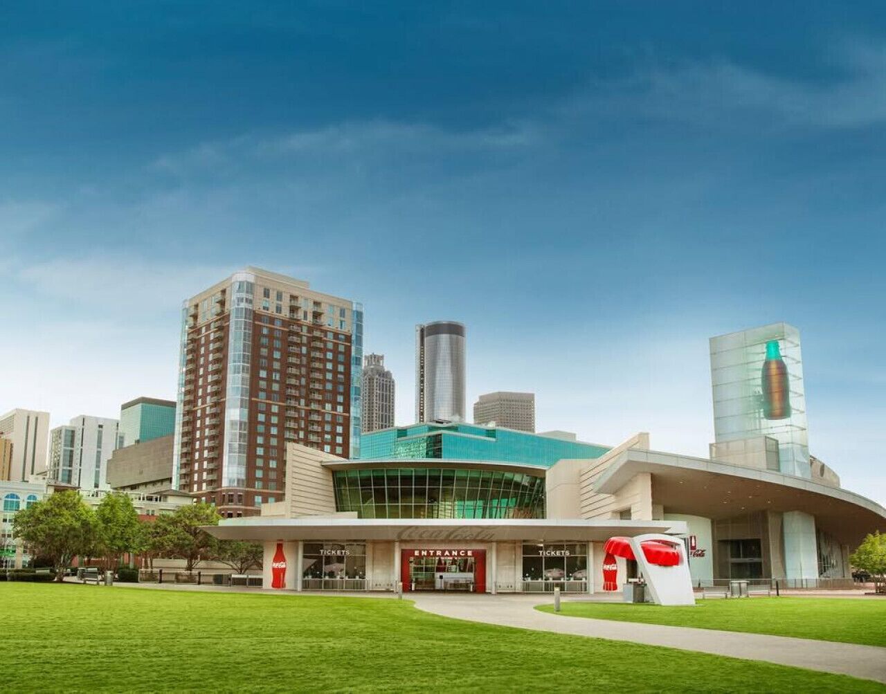 A modern building with a glass facade and a Coca-Cola logo, surrounded by a well-maintained lawn and other high-rise buildings in the background.