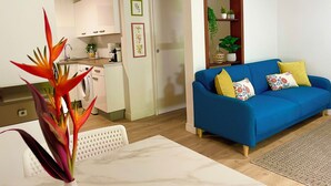 Un soggiorno con un divano blu, un tavolino bianco e una pianta. È presente anche una zona cucina con un lavello e pensili.