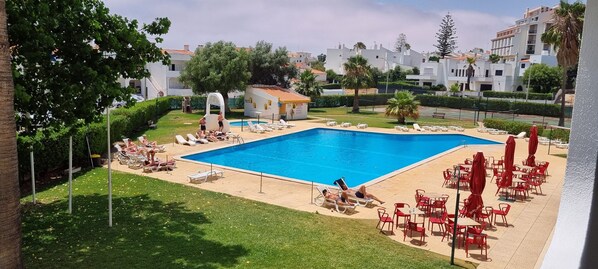 A swimming pool surrounded by lounge chairs and red umbrellas, with a small building and greenery in the background.