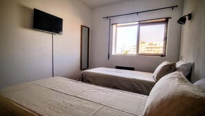 A small, simple room with two beds, a window, and a mounted TV. - Comfortable apartment in a hotel residence, Chill and festive atmosphere (Albufeira)