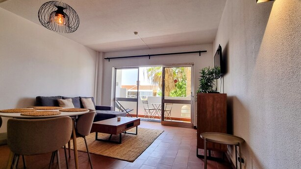 A modern living room with a sofa, coffee table, and chairs. There is a dining area with a table and chairs. A television is mounted on the wall, and a plant is on a wooden stand. - Comfortable apartment in a hotel residence, Chill and festive atmosphere (Albufeira)