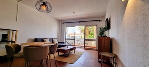 A modern living room with a sofa, coffee table, and chairs. There is a dining area with a table and chairs. A television is mounted on the wall, and a plant is on a wooden stand.