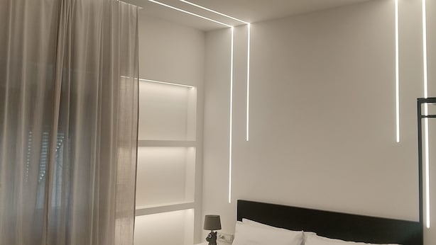 Ein modernes Hotelzimmer mit einem ordentlich bezogenen Bett, einem Nachttisch mit Lampe und einem großen Fenster mit leichtem Vorhang.