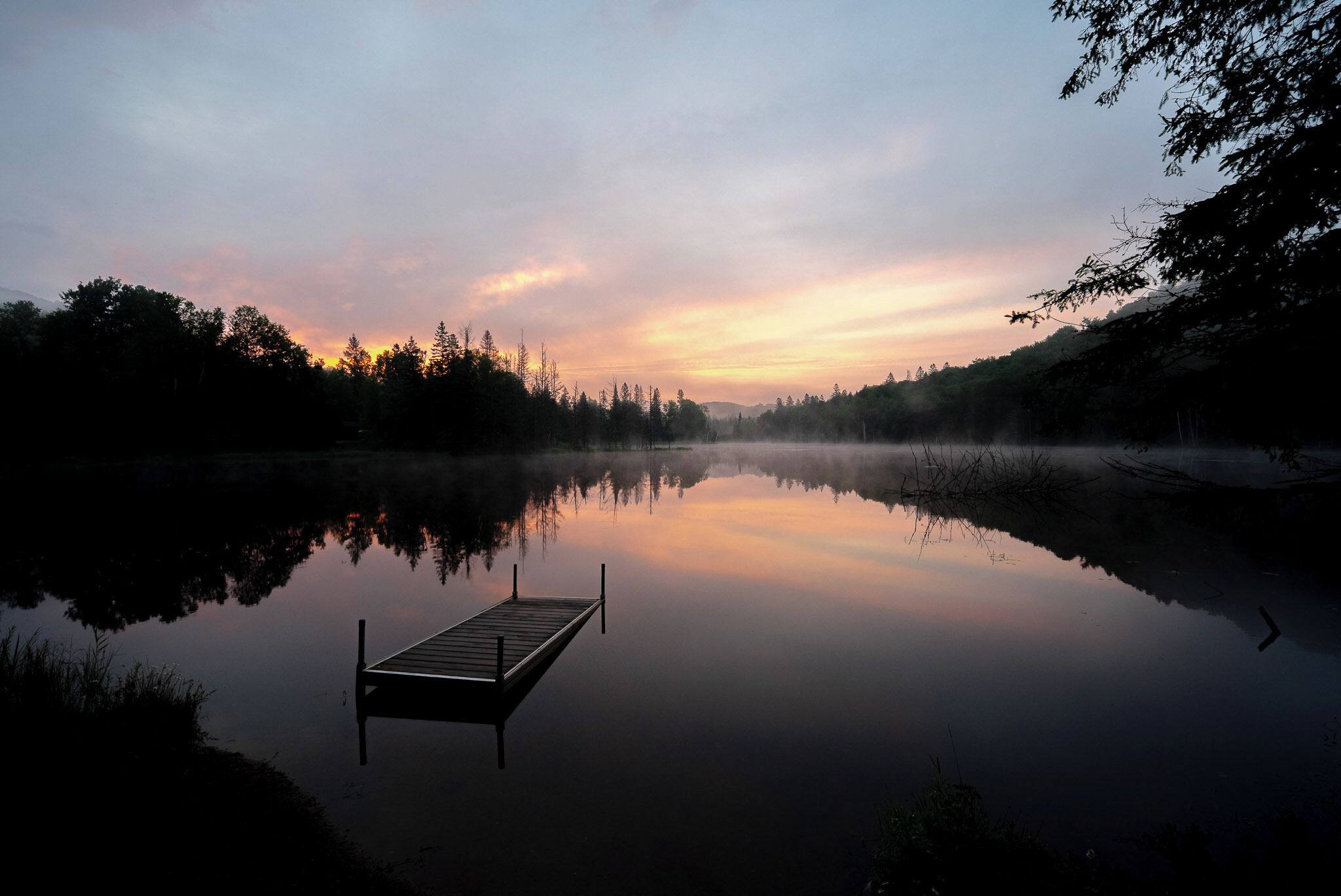 Un lago tranquilo con un muelle de madera que se adentra en el agua, rodeado de árboles y un cielo despejado.
