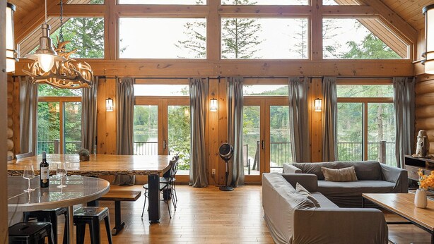 Interior espacioso de cabaña con techo alto, pisos de madera, zona de comedor, sofá y ventanas amplias que ofrecen vistas al exterior.