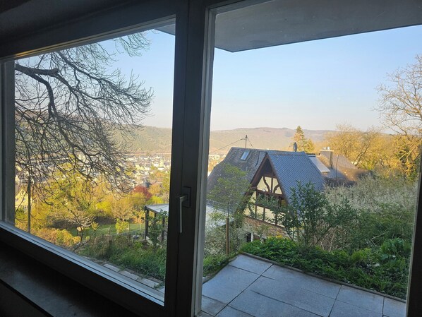 A view from a window overlooking a house with a sloped roof and solar panels, surrounded by trees and a distant town.