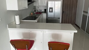 A modern kitchen with white cabinets, a stainless steel refrigerator, a microwave, and a sink. There are two red bar stools with chrome bases.