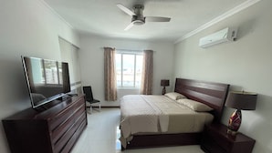 A bedroom with a bed, a TV on a dresser, a chair, a nightstand with a lamp, a ceiling fan, and a window with curtains.