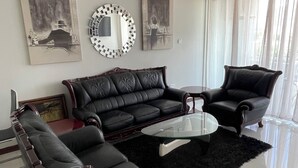A living room with a black leather sofa, a glass coffee table, and a black armchair. There is a round mirror on the wall and a painting above the sofa.