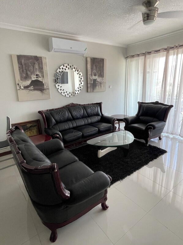 A living room with a black leather sofa, a glass coffee table, and a black armchair. There is a round mirror on the wall and a painting above the sofa.