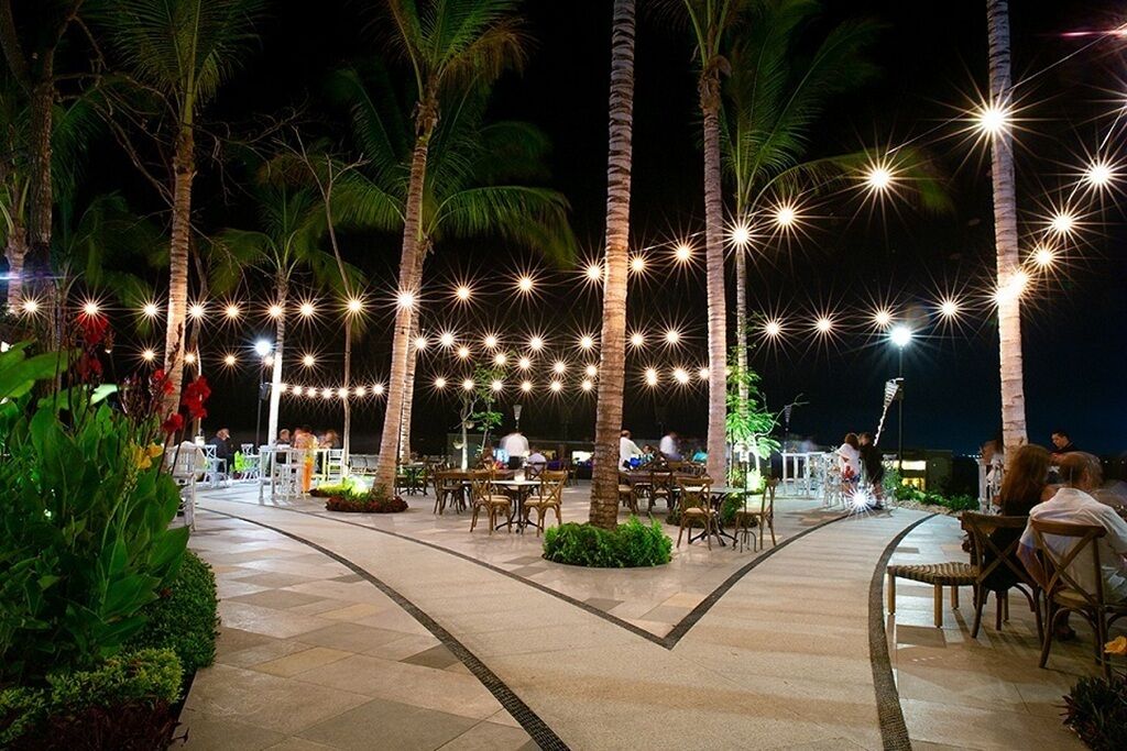An outdoor dining area with tables and chairs, surrounded by palm trees and string lights.