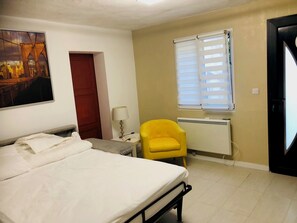 Une chambre d’hôtel avec un lit, une chaise jaune, une fenêtre avec des stores, un radiateur et une porte.