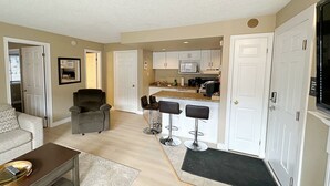 Un espacio habitable compacto con kitchenette, una zona de comedor con taburetes de bar, un sillón gris y un sofá.