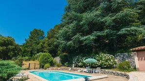 A swimming pool with lounge chairs and an umbrella, surrounded by trees and a stone wall.