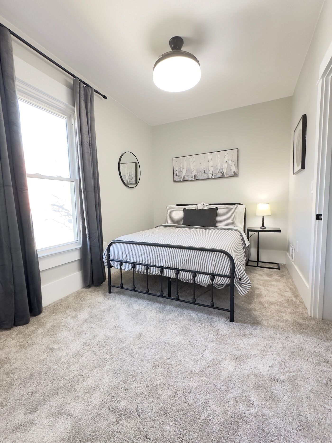 Ein Schlafzimmer mit Bett, Nachttisch, Lampe, Deckenleuchte, einem Fenster mit Vorhängen und einem an der Wand befestigten Spiegel.