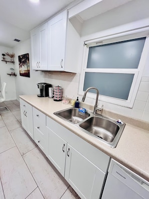 A kitchen with white cabinets, a double sink, a coffee maker, and a window with blinds.