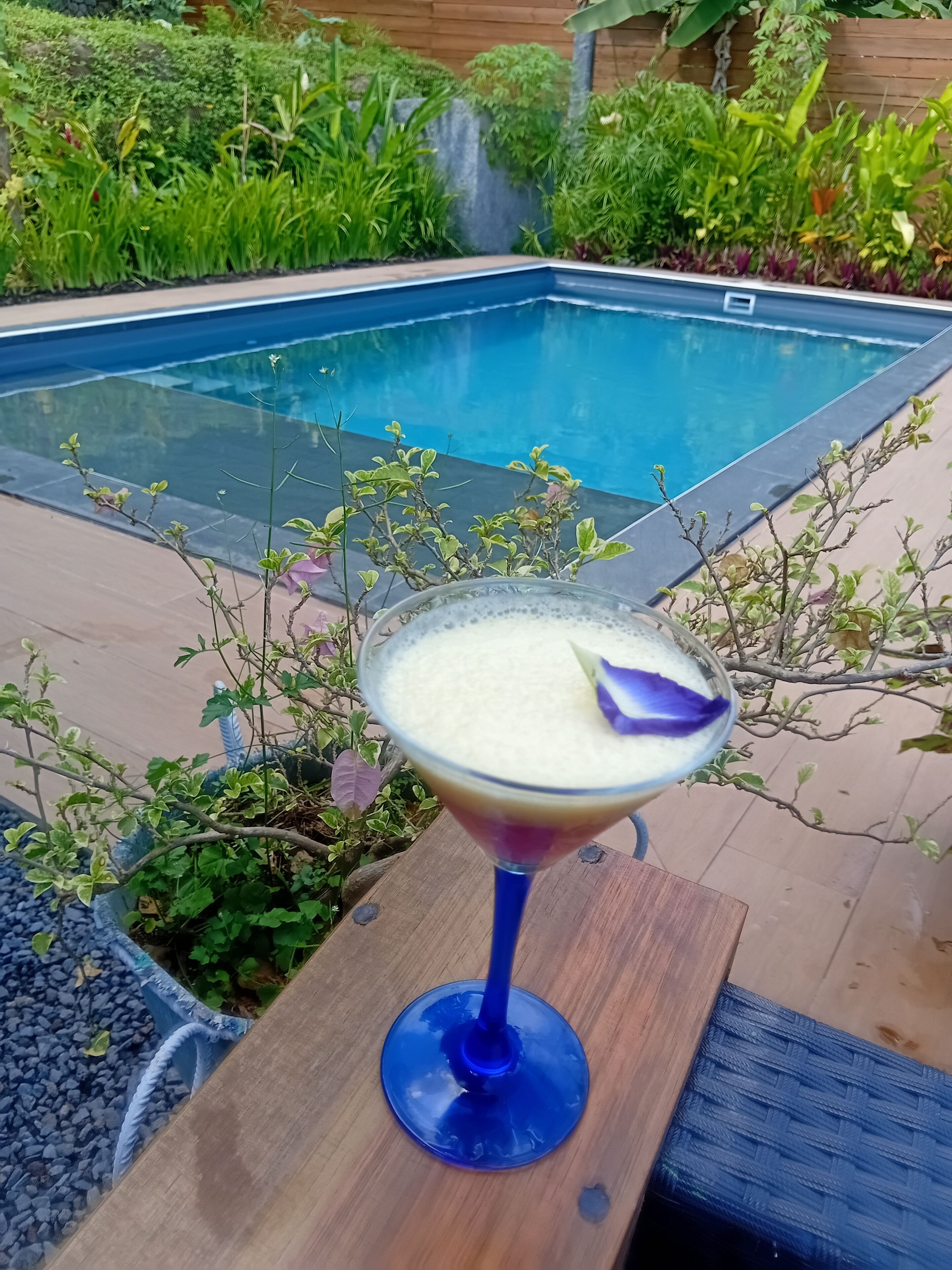 Un còctel en un got blau sobre una taula de fusta prop d'una piscina.