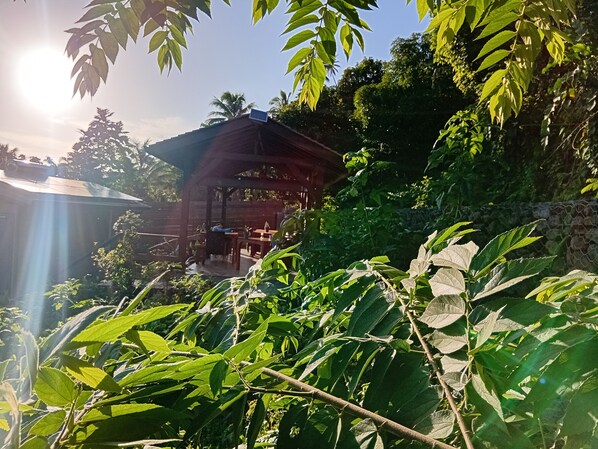 Un espacio de estar al aire libre cubierto, con mesas y sillas, rodeado de abundante vegetación y un muro de piedra.